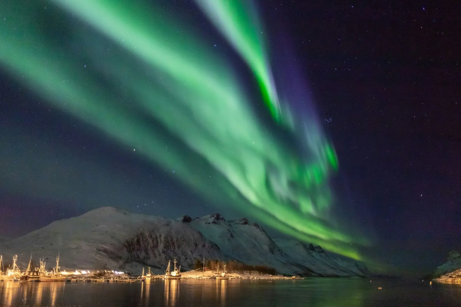 Green northern lights over snow-covered mountains and fishing boats at Ersfjord, Tromsø, Norway